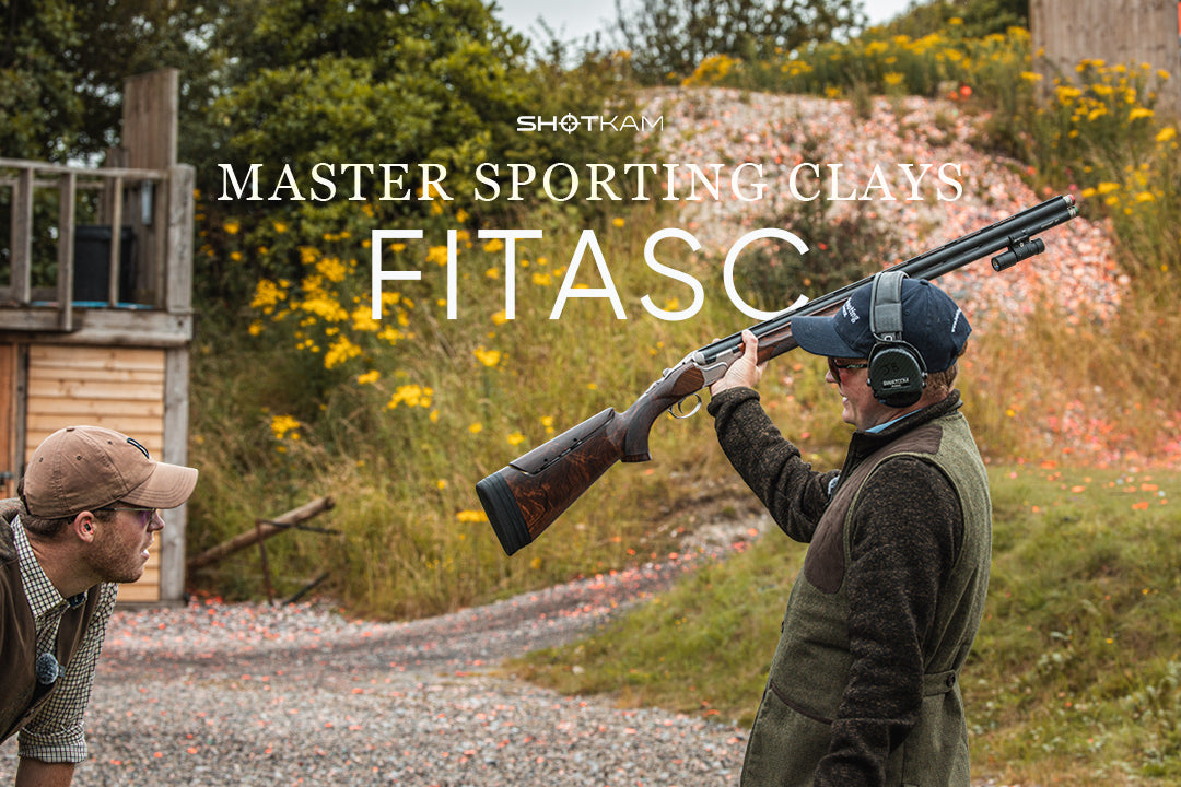 Sporting clays shooter smiling and holding shotgun overhead in grassy range, with ShotKam camera on barrel.