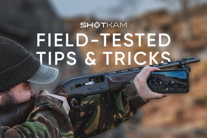 Waterfowl hunter aiming a shotgun fitted with a ShotKam camera, viewing recorded shooting footage on a mobile device.